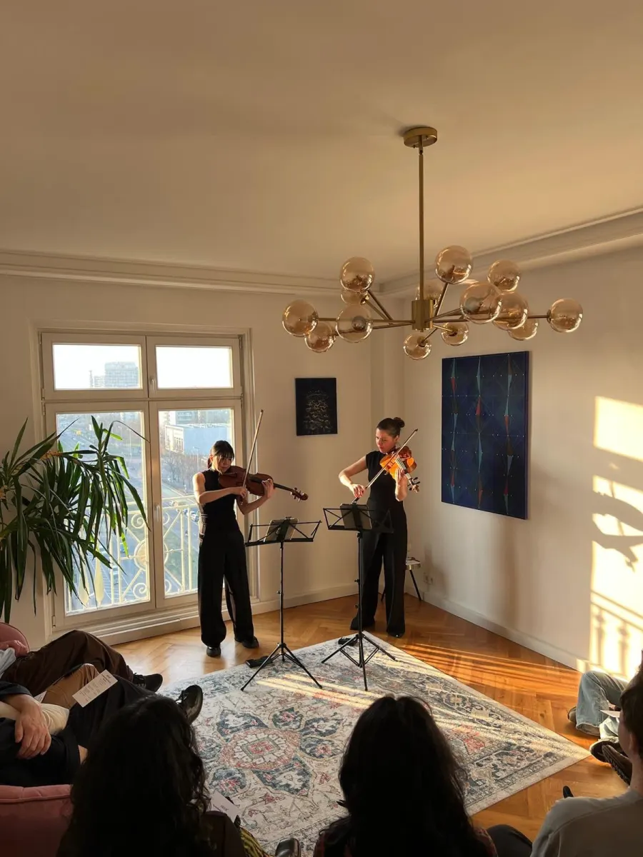 Two string players performing in a sunlit salon while the audience sits on the floor