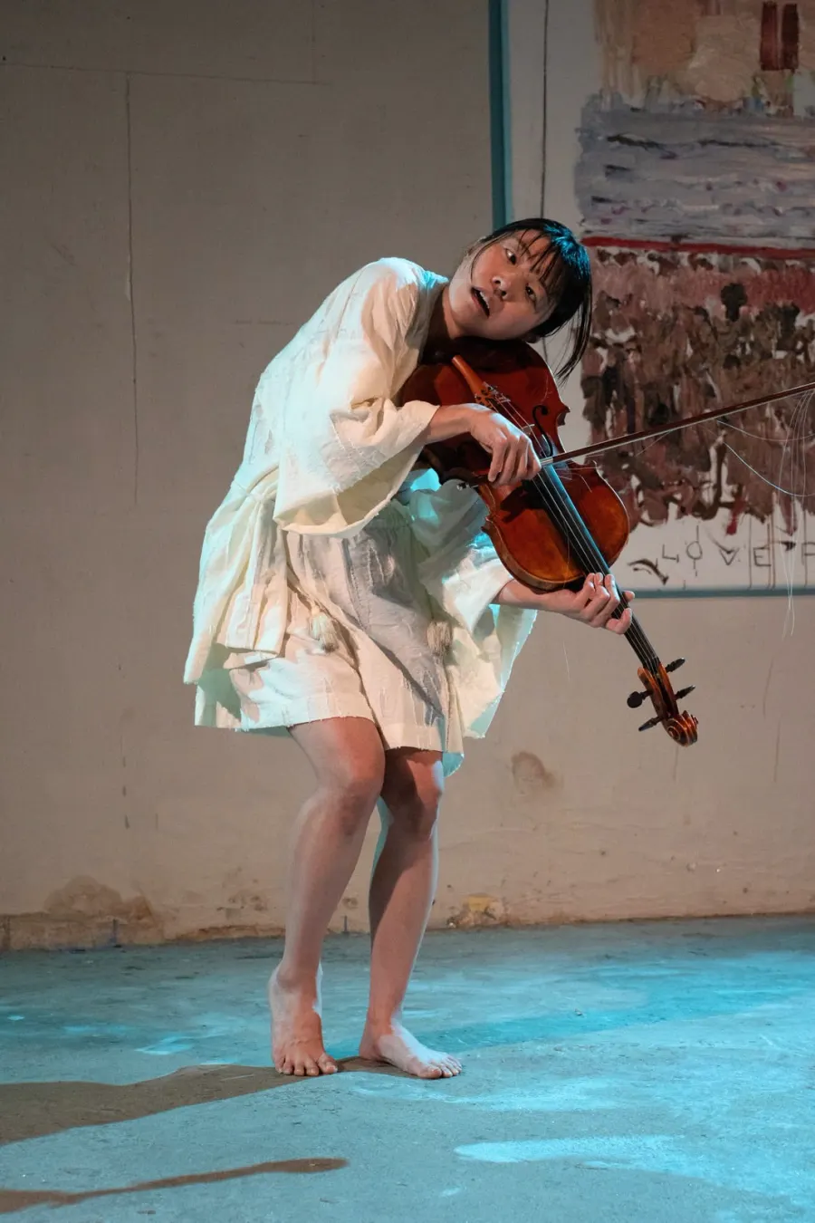 Heri Shin, barefoot in a white dress, bent forward over her viola on a pale blue floor